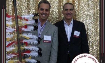 Two men standing with feathered spear in front of a gold curtain. 2019. A round seal in the corner of the image says Florida State University Jim Moran Institute Seminole 100.