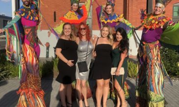 Four women in formal wear pose with four brightly costumed stilt walkers in an outdoor space.