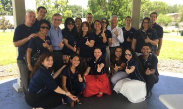 A large, happy group of people stand in an outdoor space, smiling and giving the camera thumbs up.