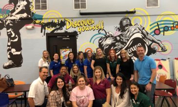 A happy group people pose in front of a wall mural depicting music and active young people and the words Discover yourself.