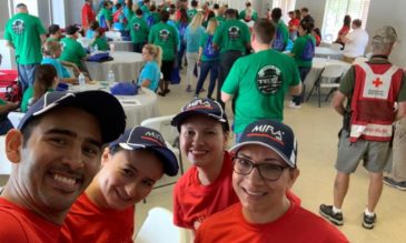 Four people in matching red t-shirts smile at the camera in front of a room full people in green and red t-shirts.