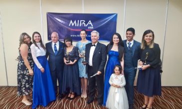 A group of adults and a child in formal wear stand in front of a banner that says MIRA USA. Four of the people are holding awards.