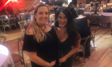 Two smiling women in black dresses stand in front of a room full of tables with red, white and black feathered centerpieces.