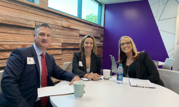 Three smiling men and women in suits sit at a round table in front of colorful, patterned walls.