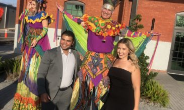 A man and a woman in formal wear pose with two brightly costumed stilt walkers in an outdoor space.