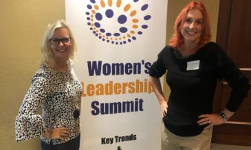 Two women standing in front of a banner that says Women's Leadership Summit Key Trends and Best Practices.