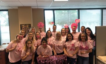 A group of women and men in pink t-shirts holding pink prop glasses, crowns, lips and hair. The t-shirts say Taylor White and show the breast cancer support ribbon inside a pink heart.