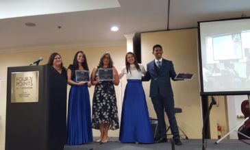 Five people in formal wear standing on a stage holding certificates. A sign on a nearby podium says Four Points by Sheraton.